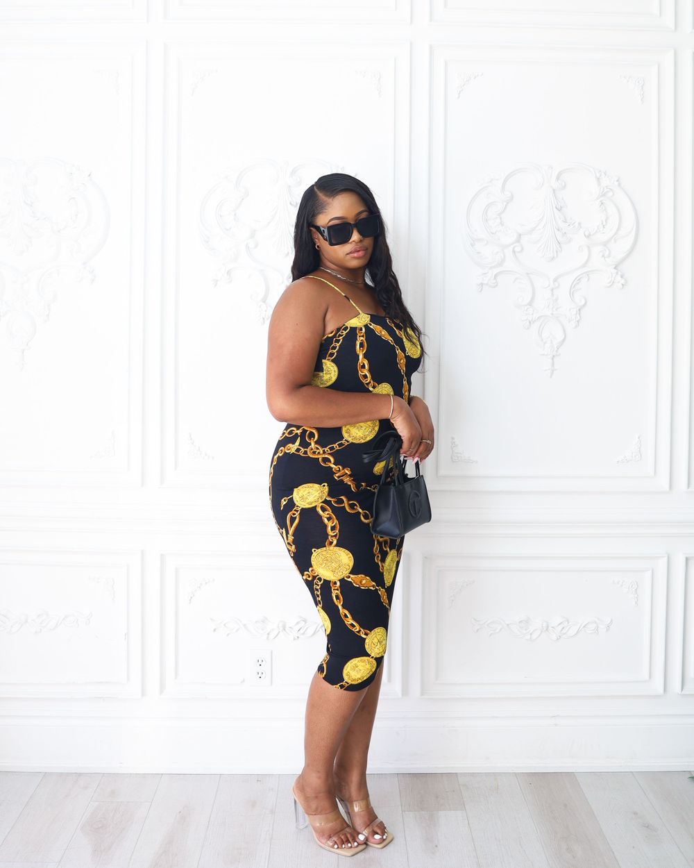 Fashion model poses in black and gold patterned dress against white ornate wall.