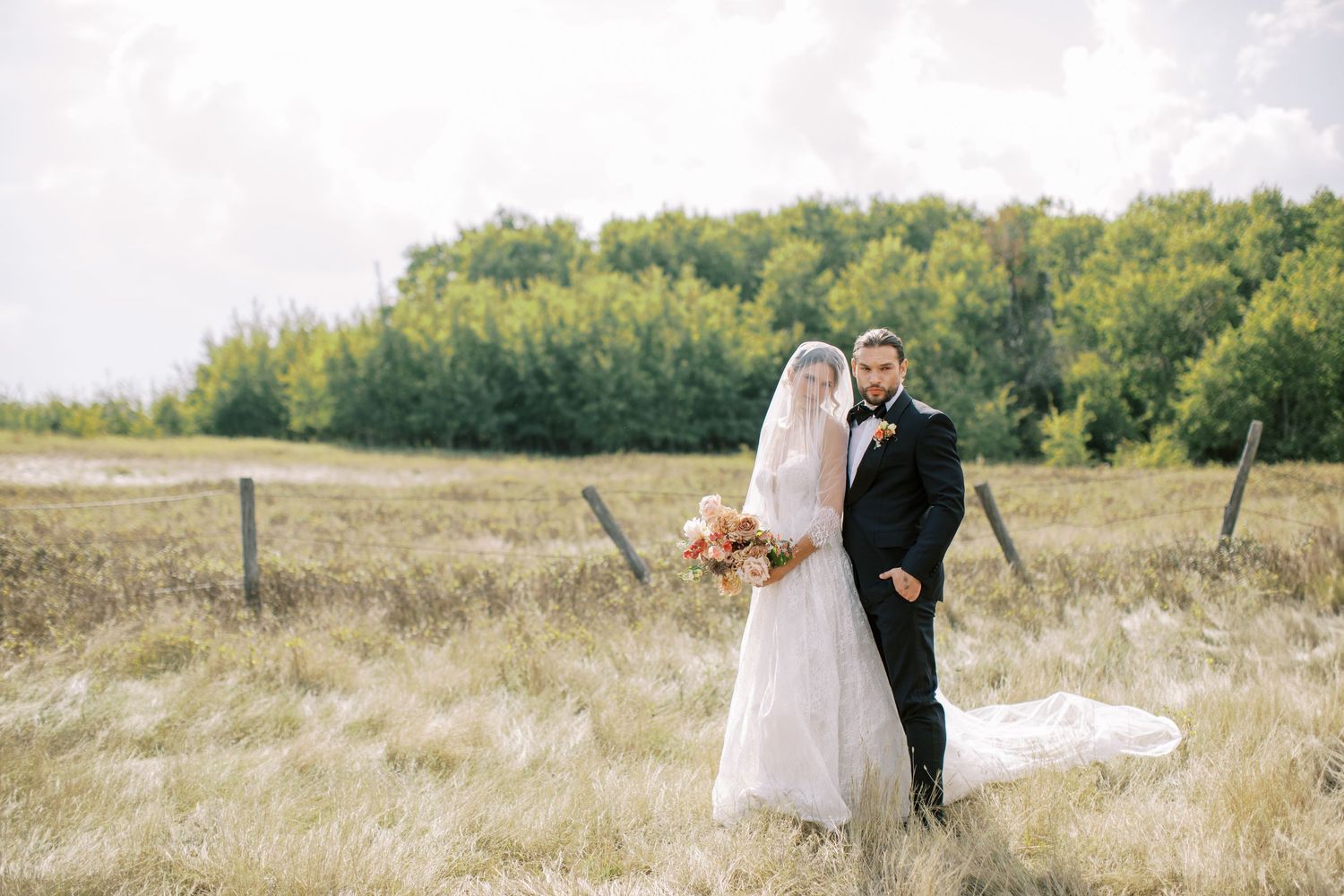 Bride in flowing white dress and groom in dark suit pose playfully in a grassy field.