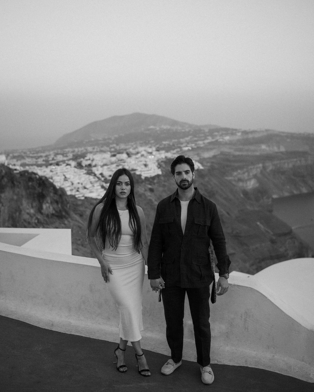Couple posing against stunning Santorini caldera view with white buildings cascading down hillside.