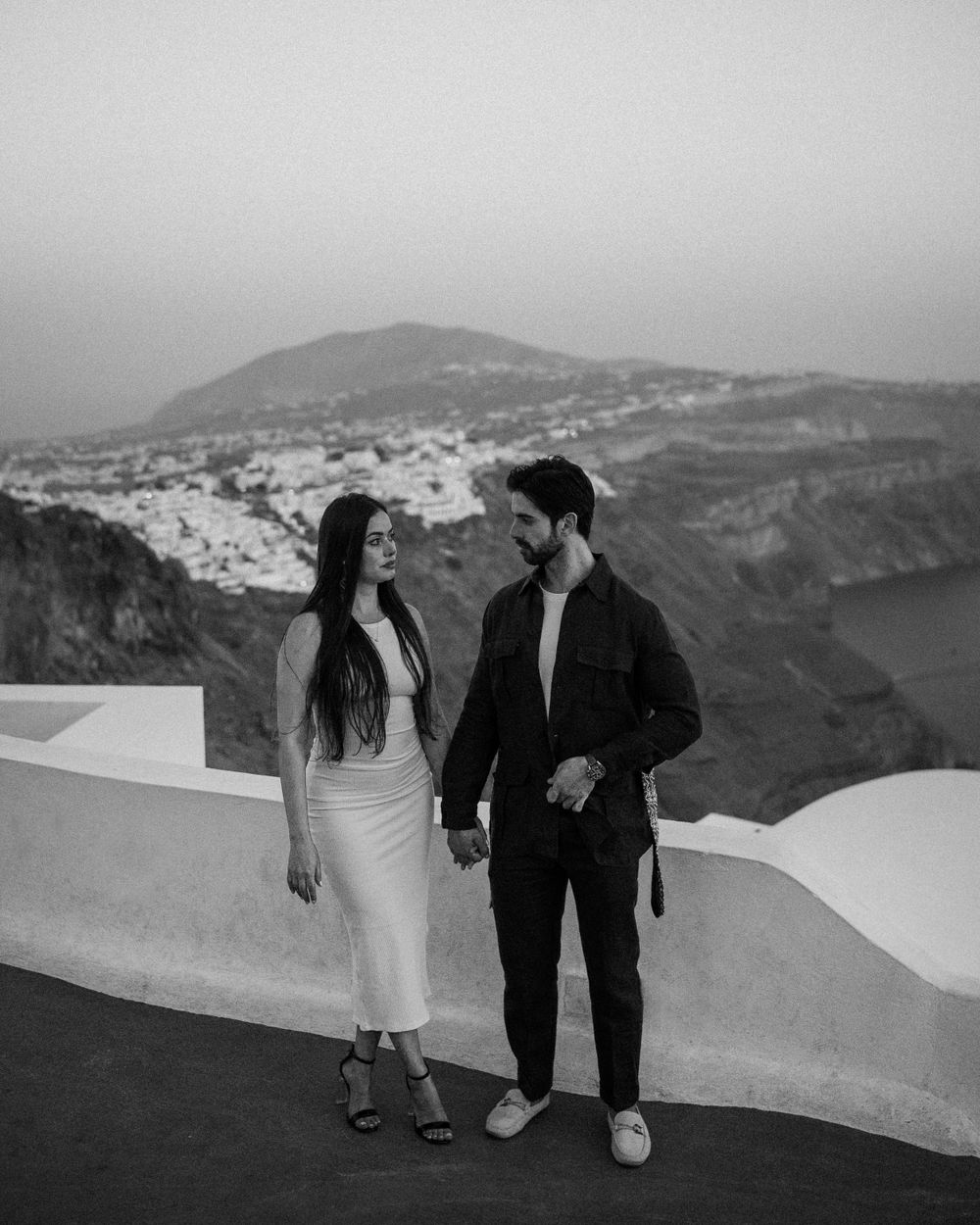 Black and white photo of couple standing on white terrace overlooking Santorini's dramatic coastal landscape.