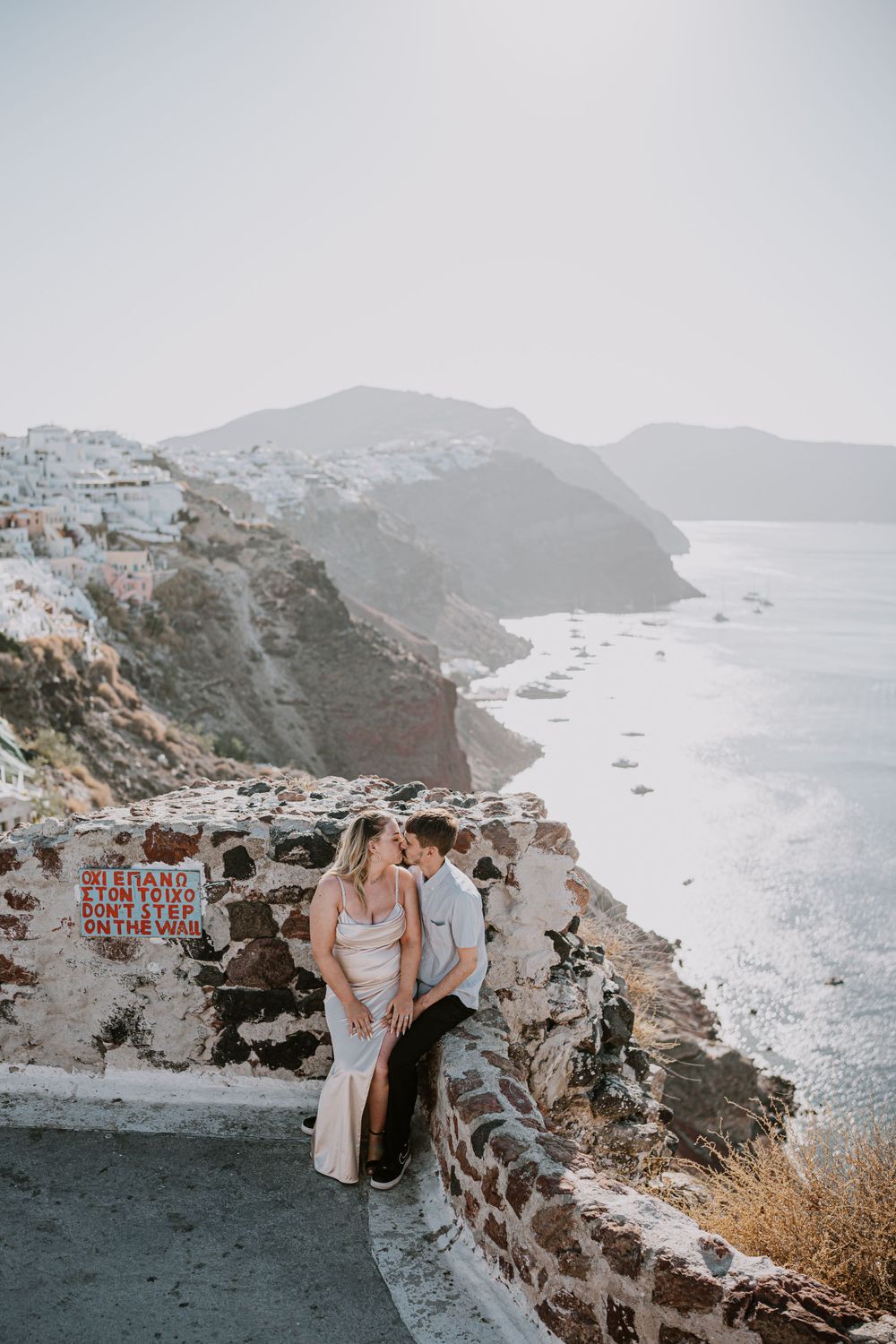 Couple sharing romantic moment on stone wall overlooking dramatic Santorini coastline.