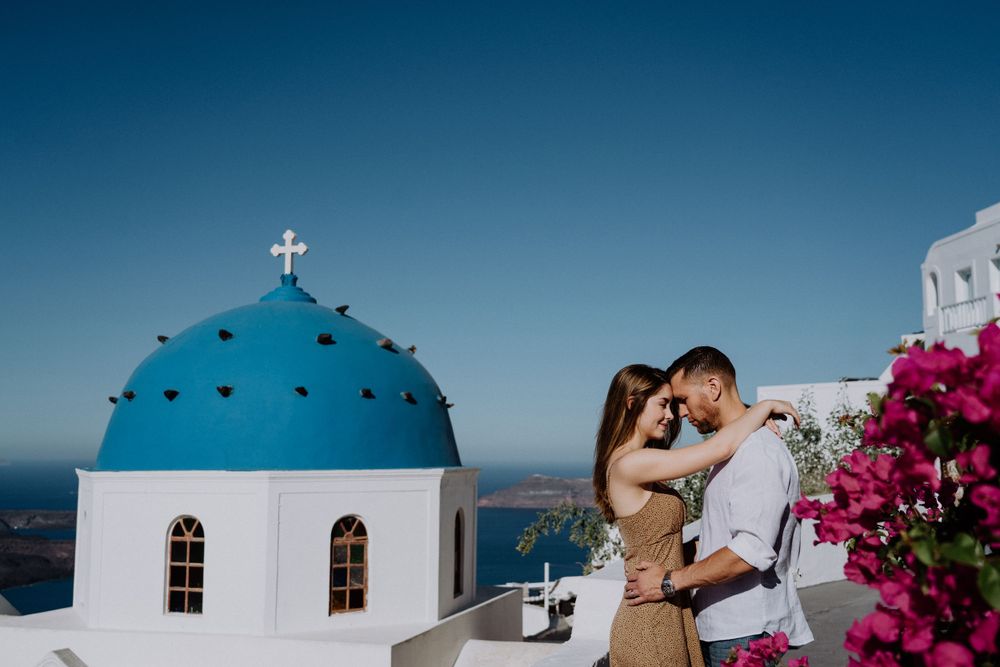 Romantic moment by blue-domed church with pink bougainvillea in Santorini.