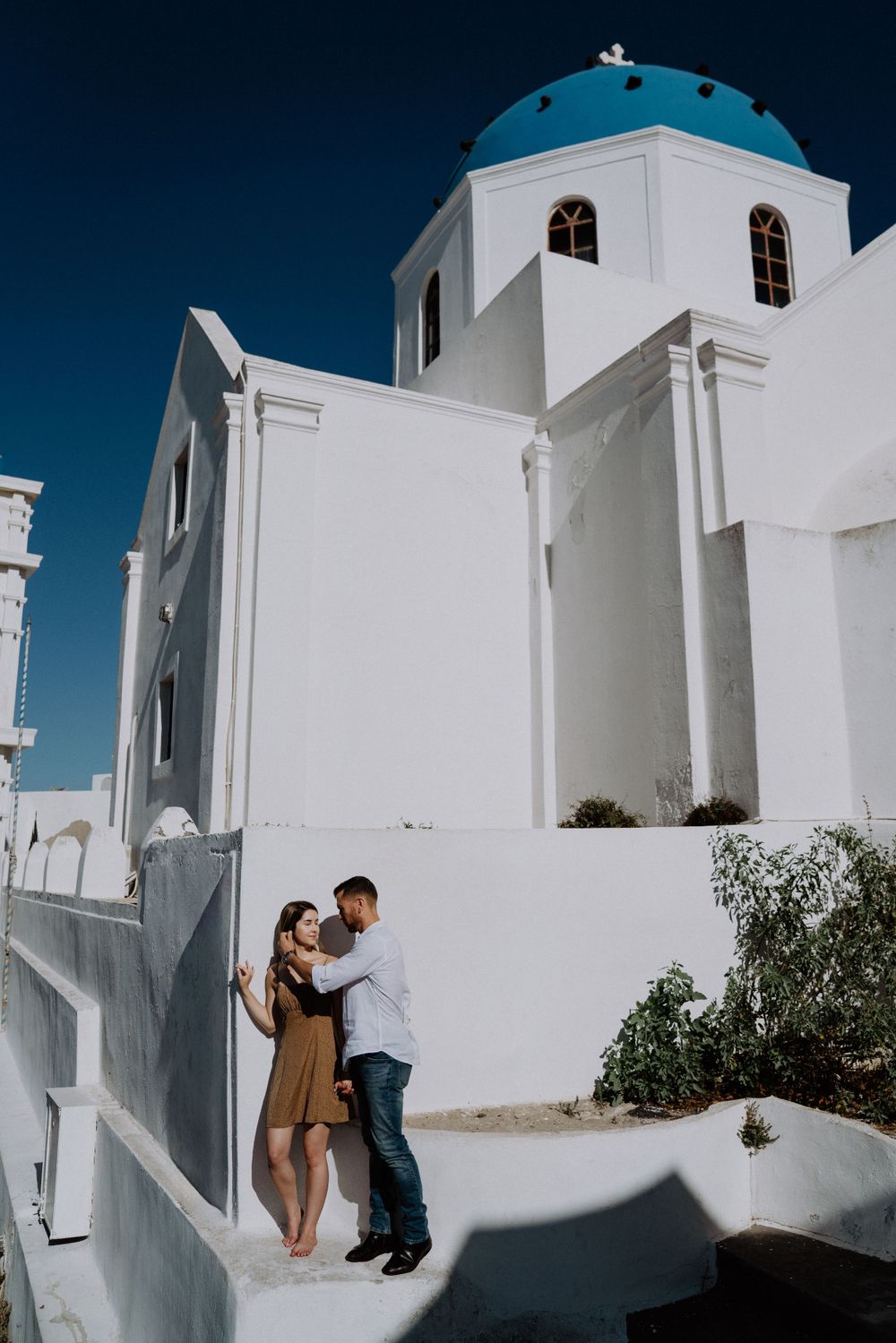 Couple embracing by traditional white Greek Orthodox church with blue dome in Santorini.