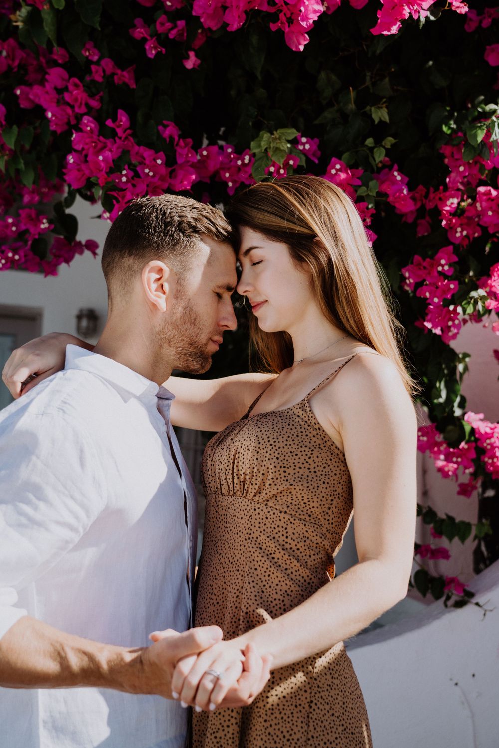 A romantic couple embraces under vibrant pink bougainvillea flowers against a white wall.