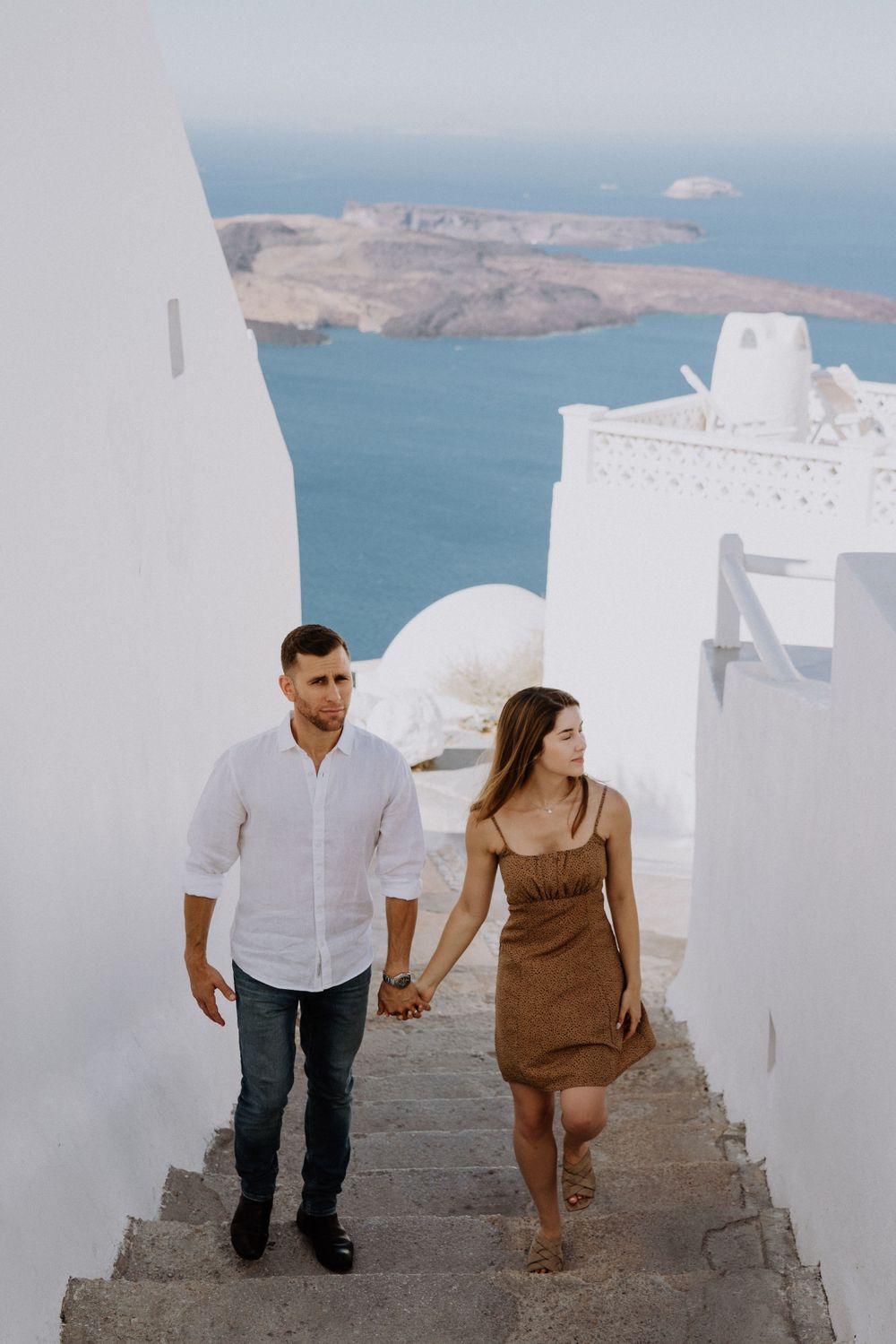 A couple walks hand in hand down white steps in Santorini with ocean views.