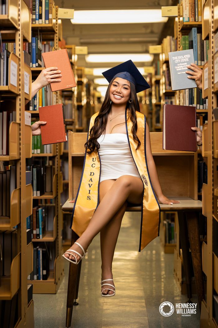Graduate in red cap poses against red backdrop wearing blue sweater and white skirt for portrait photo.