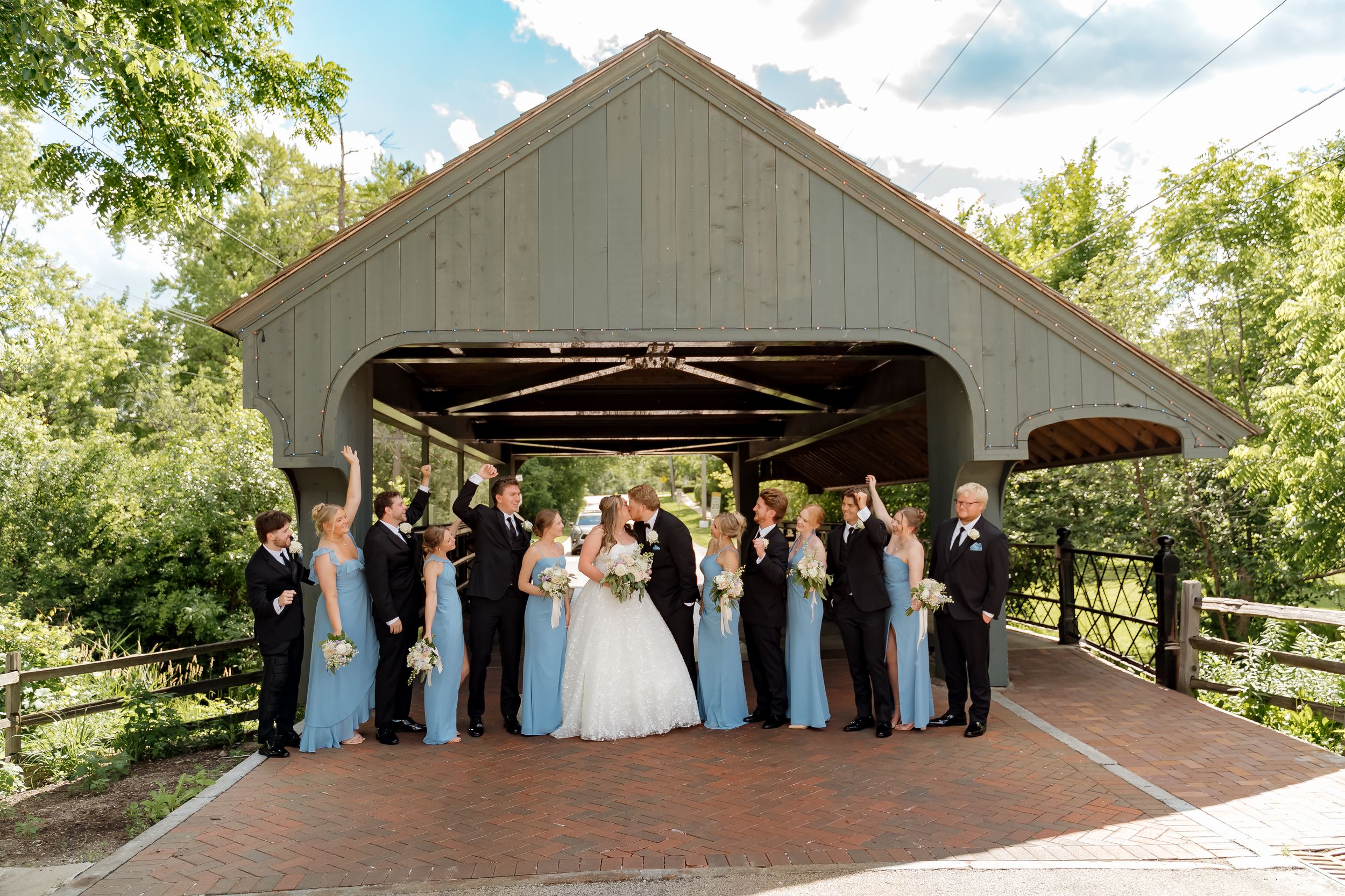 Wedding party poses before a covered bridge, bridesmaids in blue gowns and groomsmen in black suits celebrating outdoors.