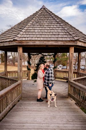 A couple stands with their dog in a wooden gazebo on a bright winter day.