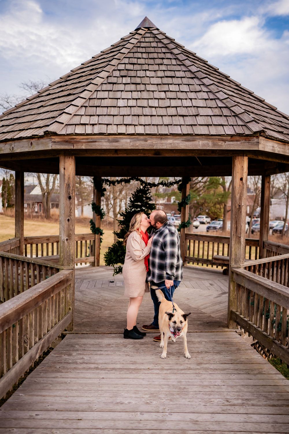 A couple stands with their dog in a wooden gazebo on a bright winter day.