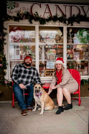 A couple sits with their dog on red chairs outside a vintage candy store decorated for Christmas.
