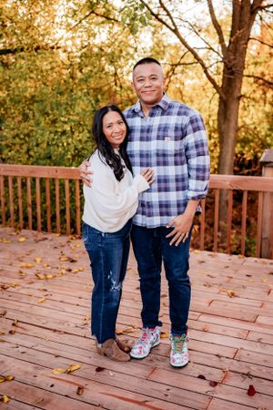 Couple in casual wear poses on wooden deck with autumn foliage in background.