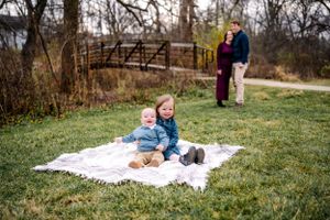 A child sits on a blanket in a park with a bridge and couple visible in background.