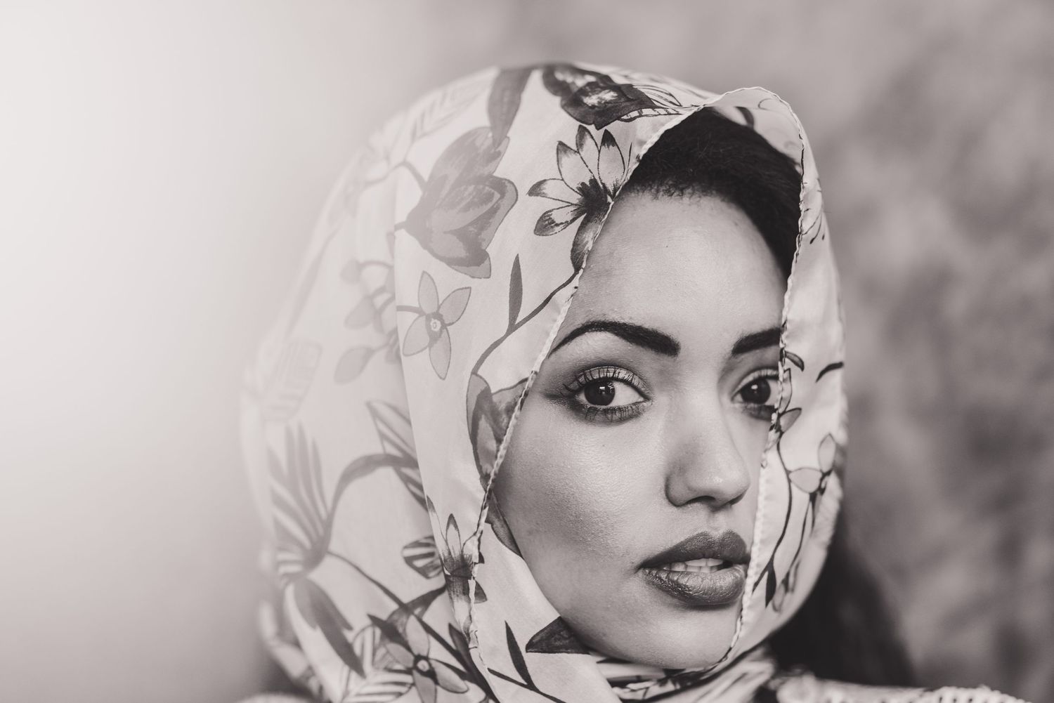 Black and white portrait of someone wearing an elegant floral patterned hijab against a blurred background.