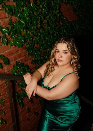 Elegant poses in an emerald green satin dress against a brick and ivy wall.