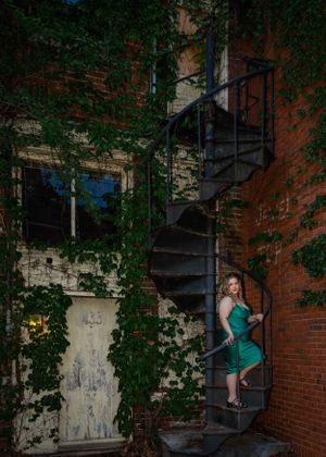 Figure in emerald dress poses on vintage spiral staircase with ivy-covered brick wall.