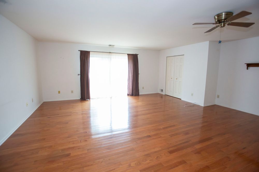 Empty living room with hardwood floors, ceiling fan, and sliding glass door with curtains.