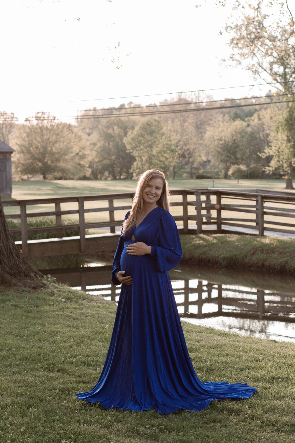 A romantic maternity photoshoot in a flowing royal blue dress by a wooden fence and pond at sunset.