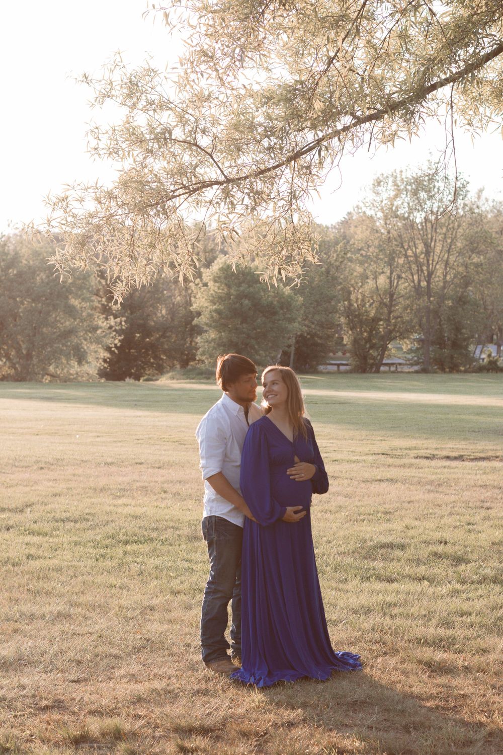 A couple shares tender moments during an outdoor maternity photoshoot in a grassy field at golden hour.