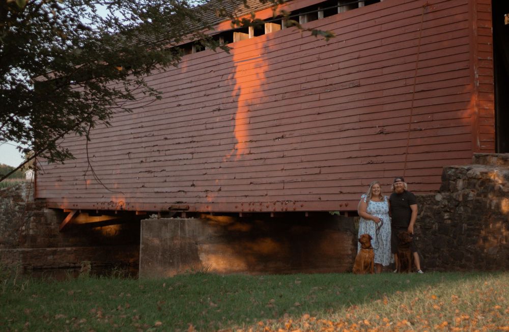 Couple posing by a rustic red covered bridge at sunset.