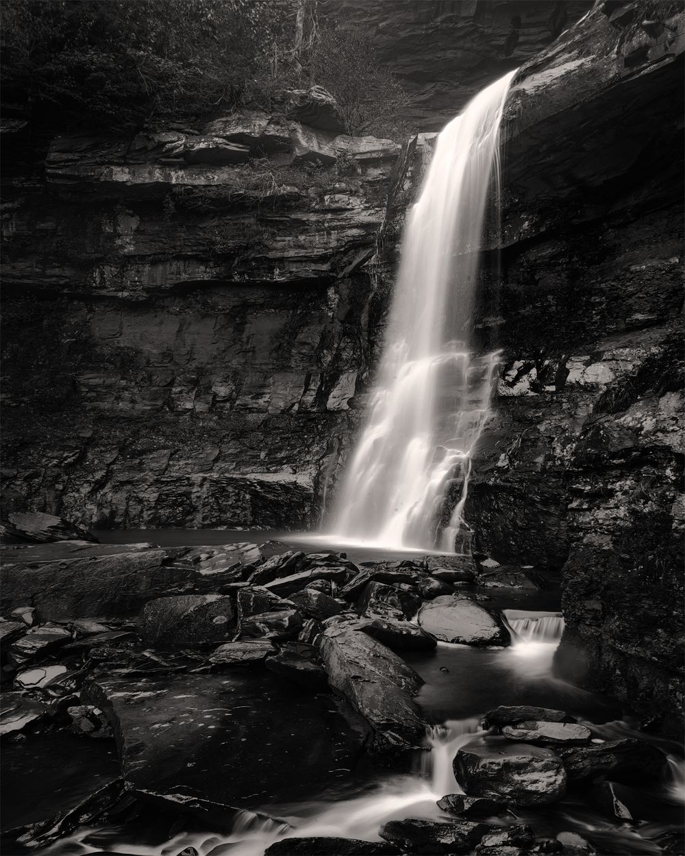 Dramatic black and white waterfall cascading down layered rock face with scattered boulders below.