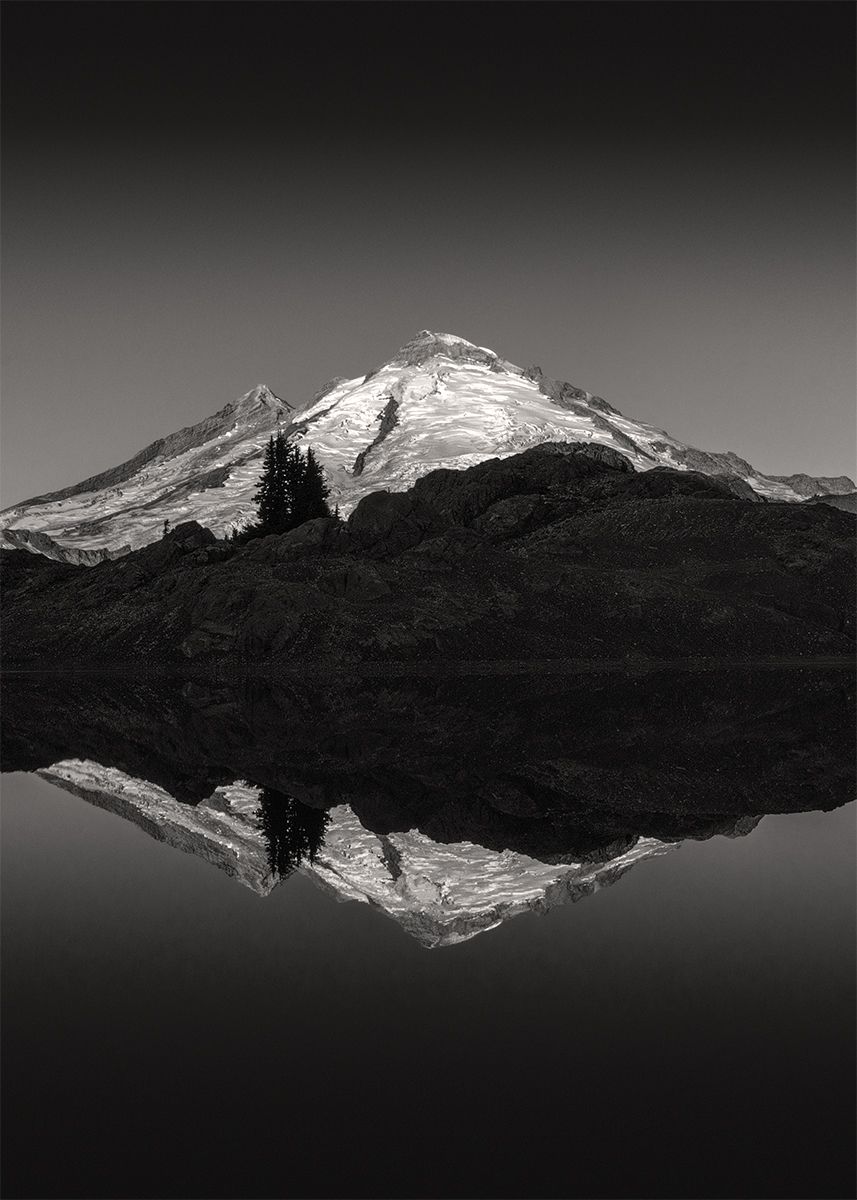 A snow-capped mountain peak reflects perfectly in a still lake with dark forested slopes in black and white.