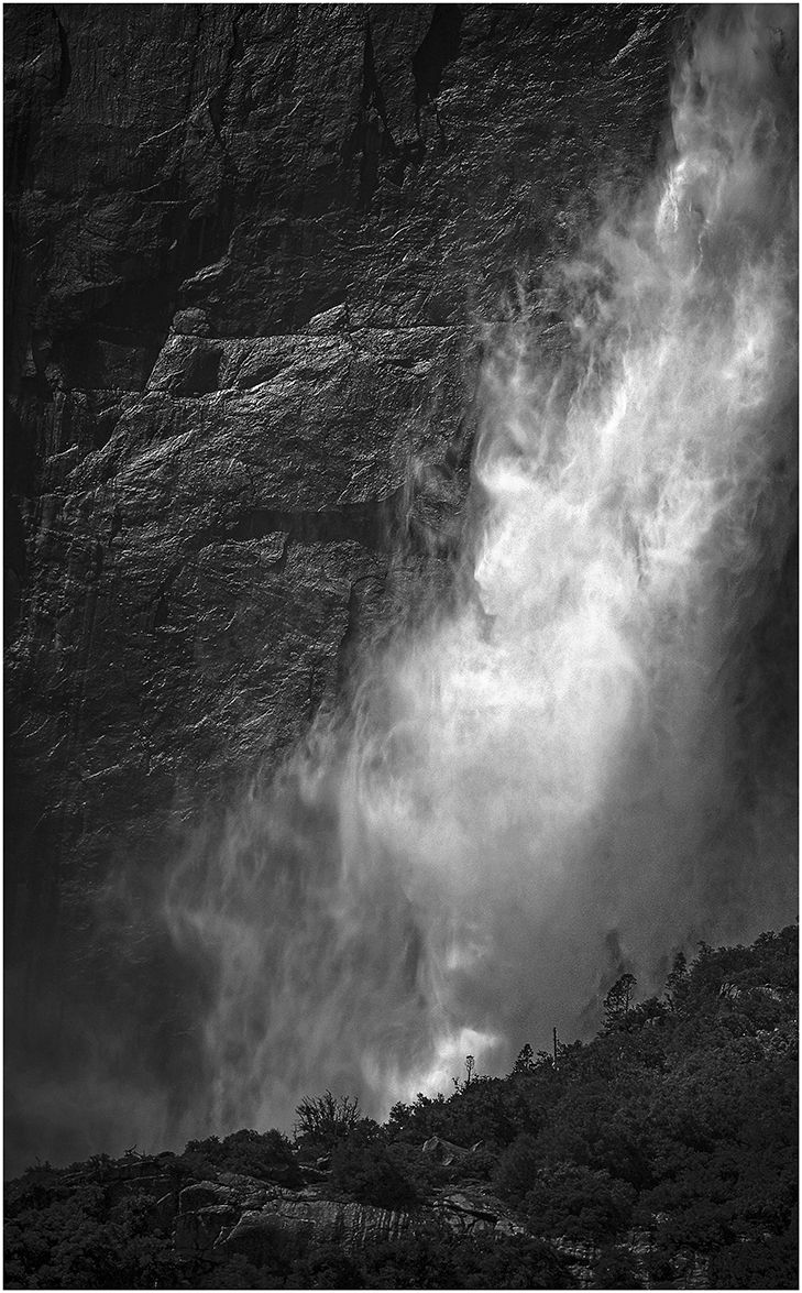 Dramatic black and white waterfall cascades down a steep rock face creating misty ethereal effects against dark cliffs.
