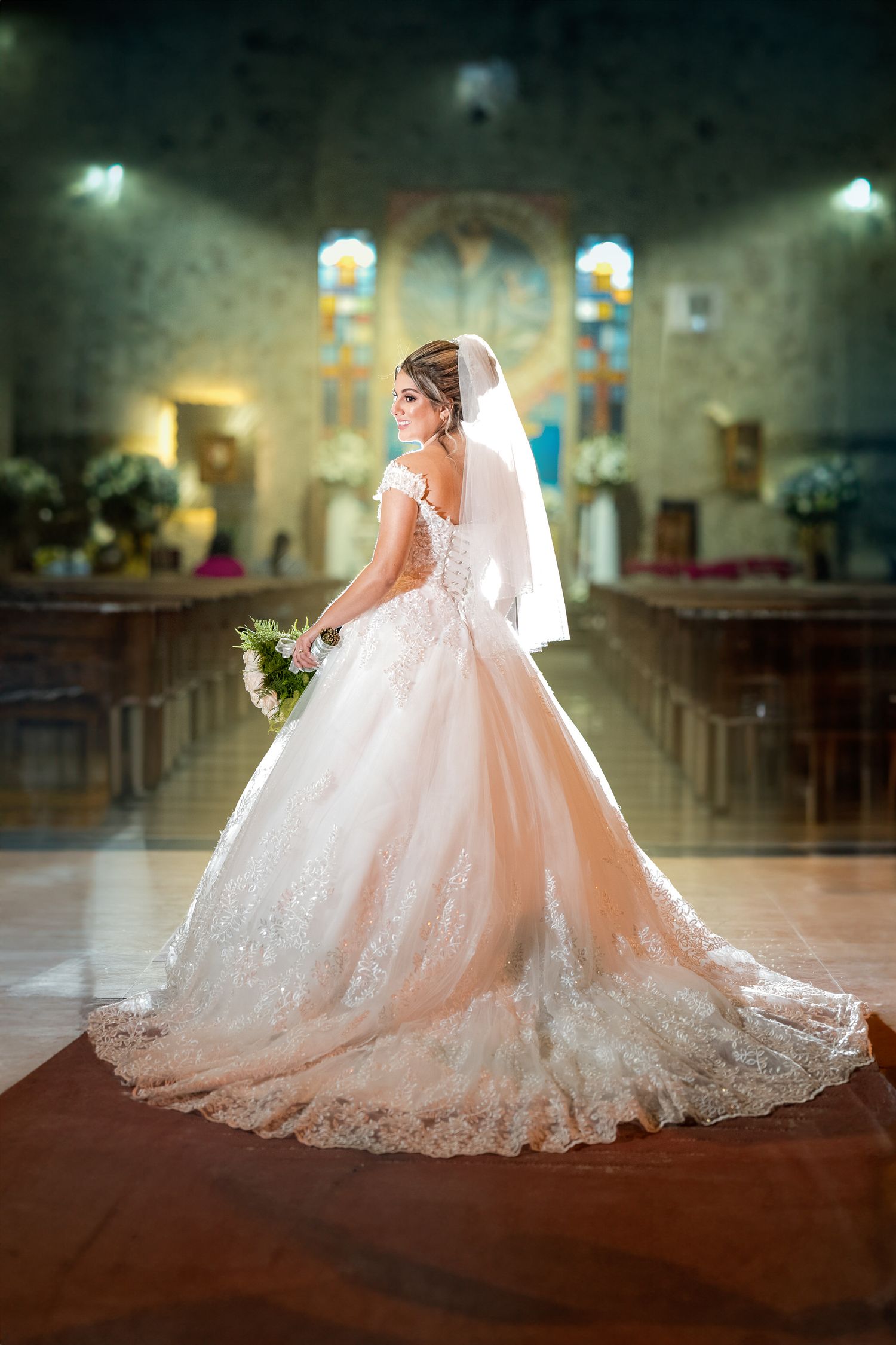 Elegant bride in a lace wedding gown stands in a beautifully lit church, holding a bouquet and gazing back with a radiant smile.