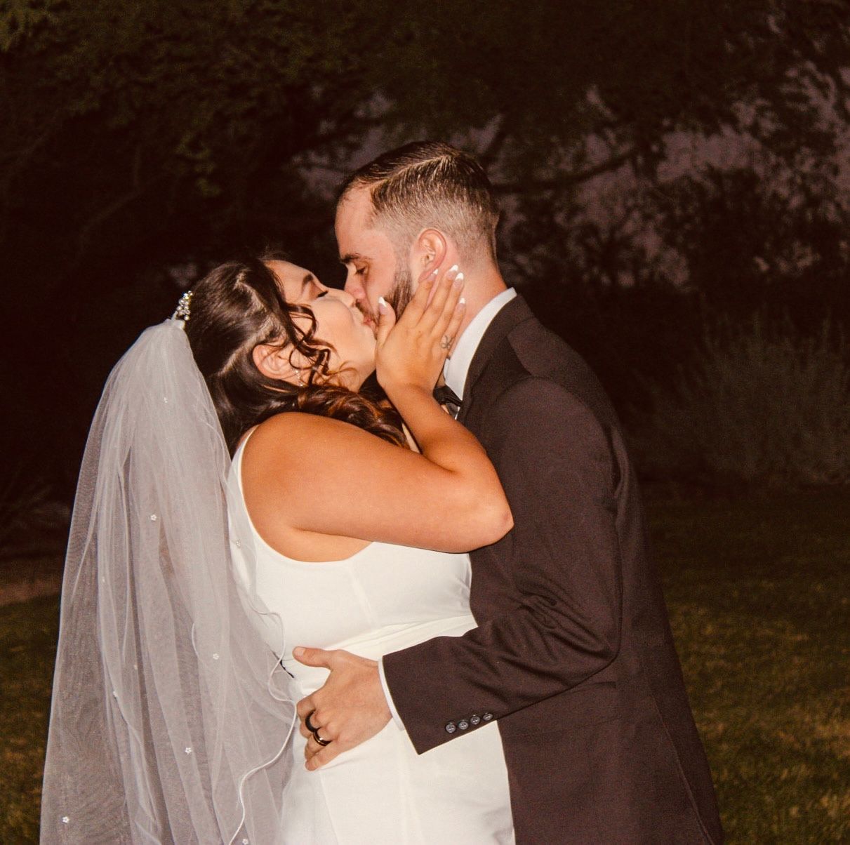 Newlyweds share a passionate kiss outdoors at night, bride wearing veil in wedding gown.