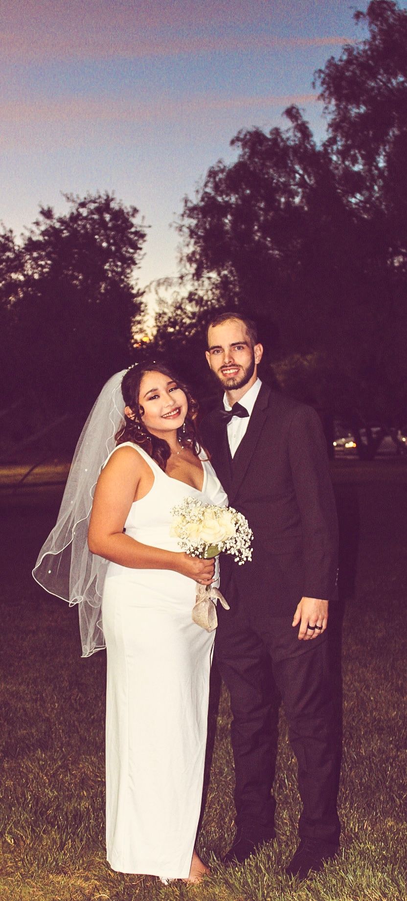 Bride holding white bouquet and groom in black tuxedo pose together at sunset outdoor wedding.