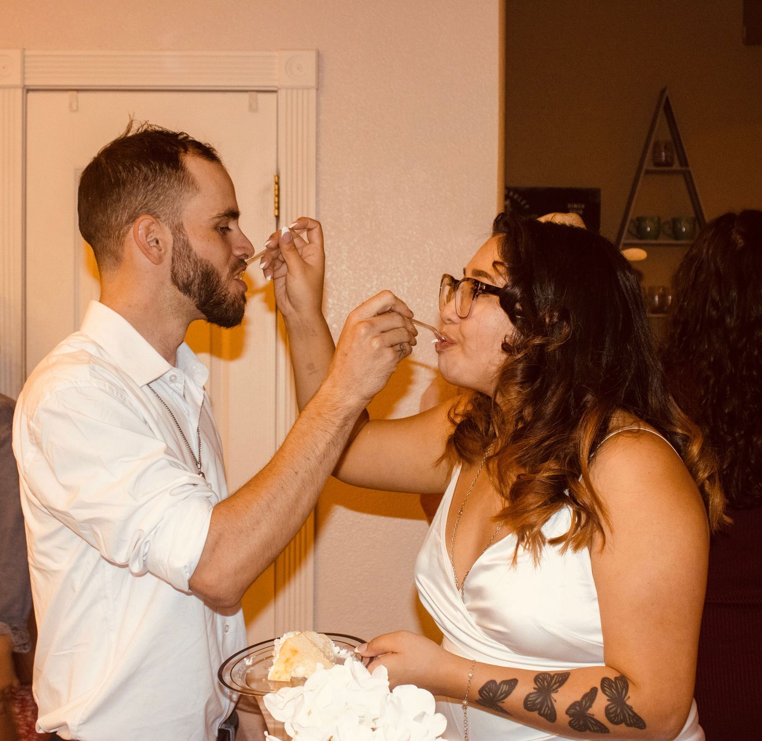 Newlyweds share fun cake-feeding moment indoors, bride with tattoo and groom in white shirt laughing.