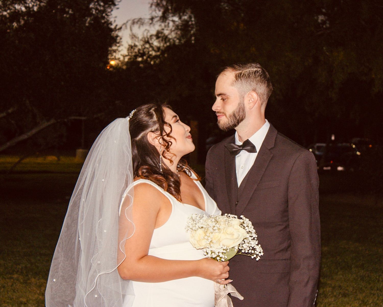Bride and groom share a romantic moment outdoors at dusk, bride holding white bouquet and wearing veil.