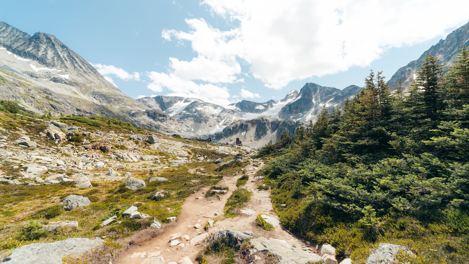 A winding dirt trail leads through rocky alpine terrain with snow-capped mountains and evergreen trees in the background.
