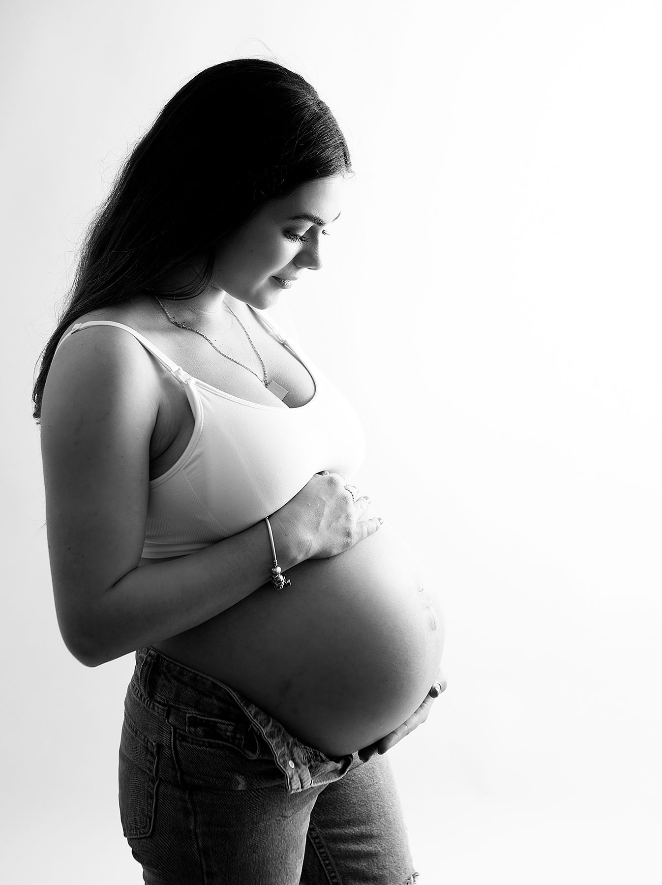 Black and white profile portrait of an expectant mother in a white tank top tenderly cradling her pregnant belly. During a shoot in Bexley