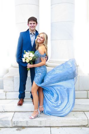 Couple in formal attire pose on white steps, with her wearing a flowing blue gown and him in a navy suit.