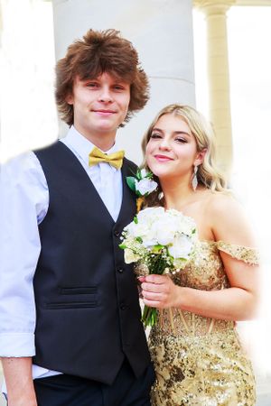 Formal couple posing by white pillars, with her in gold dress and him wearing black vest with bowtie.