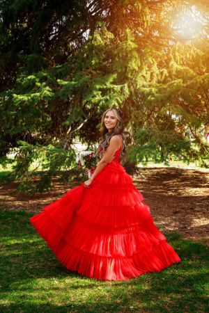 Young woman in red ruffled ball gown twirls in garden with sunlit trees.