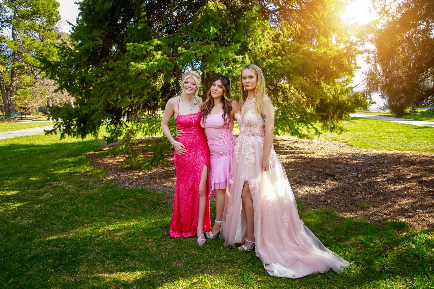 Three friends in formal prom dresses pose together in a sunlit garden setting.