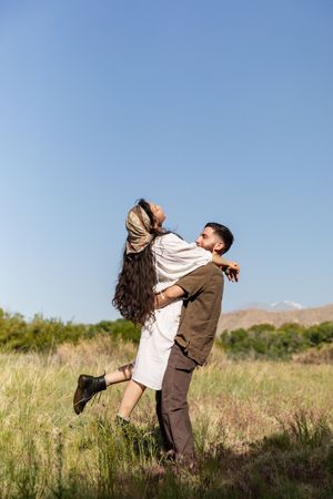 Joyful couple embraces and spins in a grassy field against blue sky and mountains on a sunny day in Jacksonville NC.