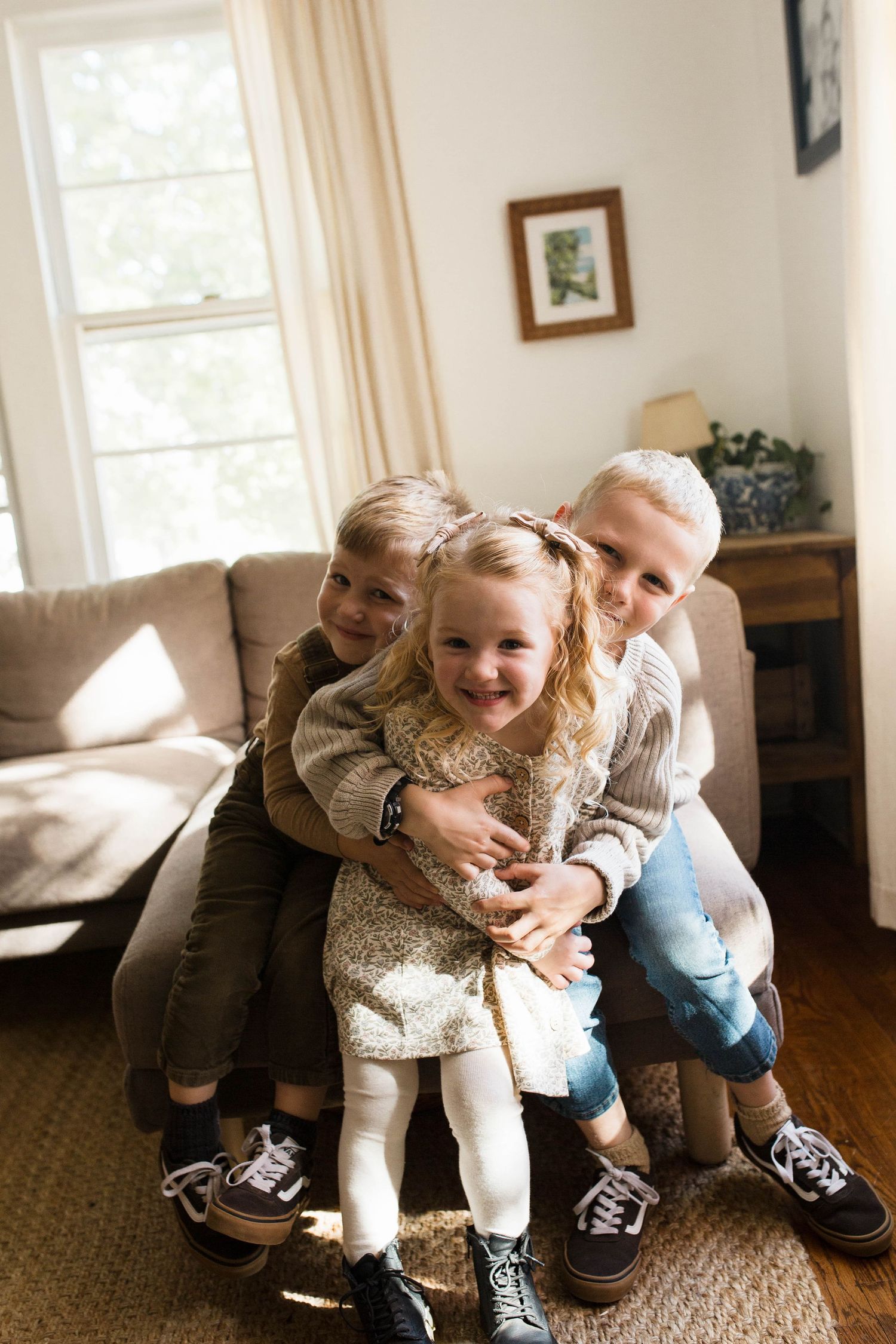 Playful moment captured by B. Logsdon Photography in sunny living room as children share laughter on brown couch by window.