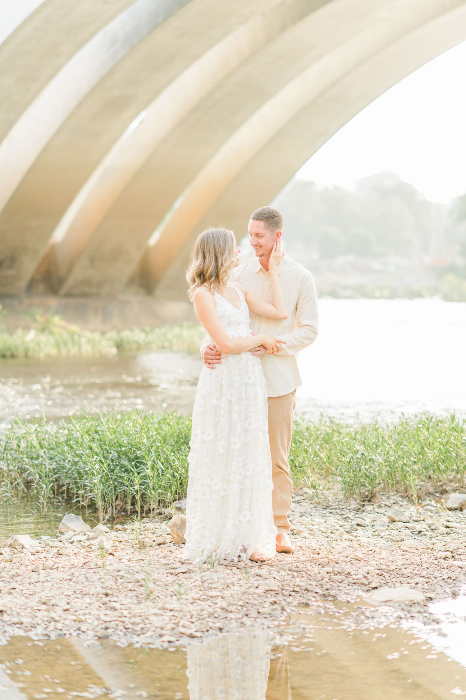 A couple shares a tender moment near a river under a concrete arch bridge in soft golden light.