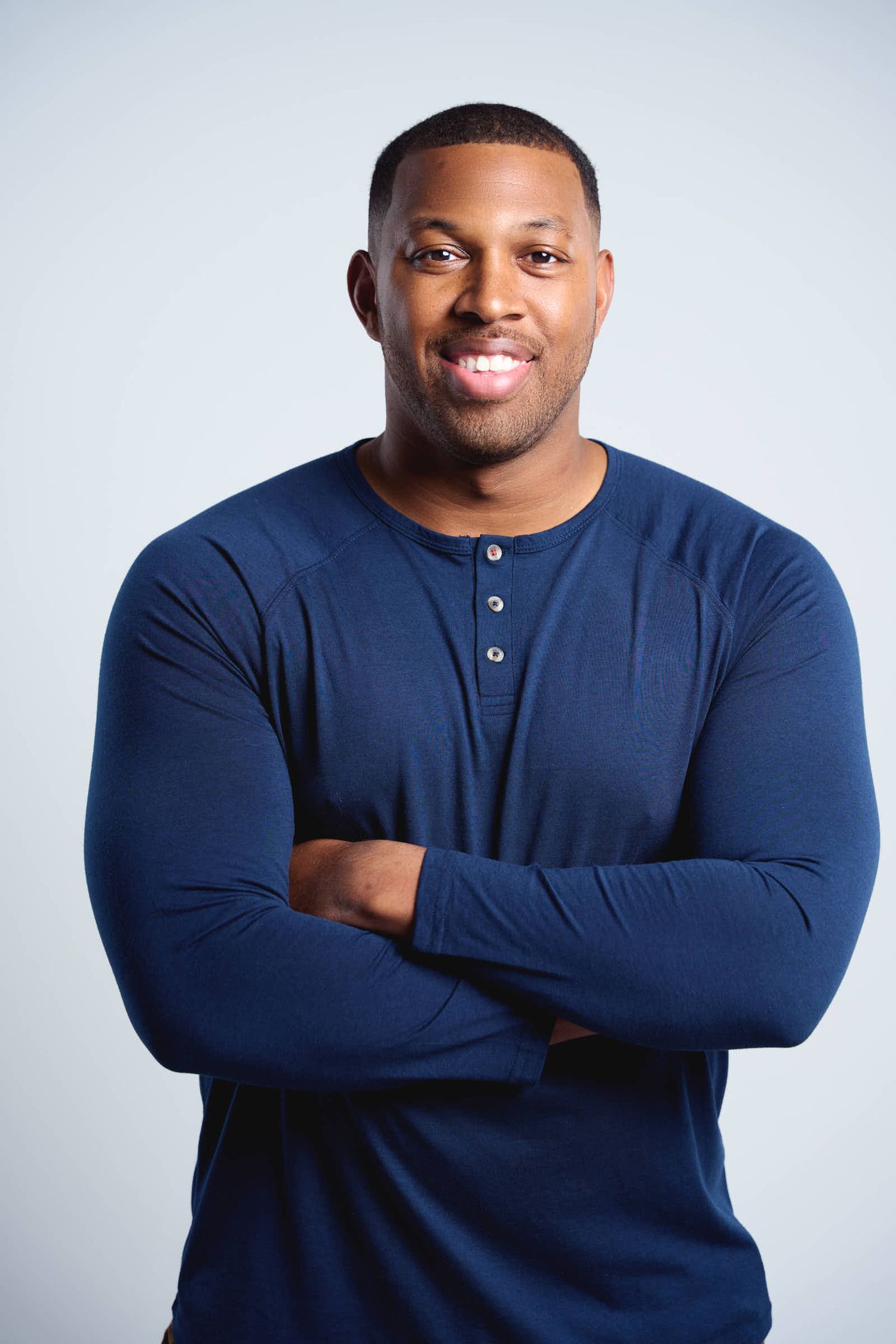 Smiling man in navy henley shirt with crossed arms poses against light gray background.