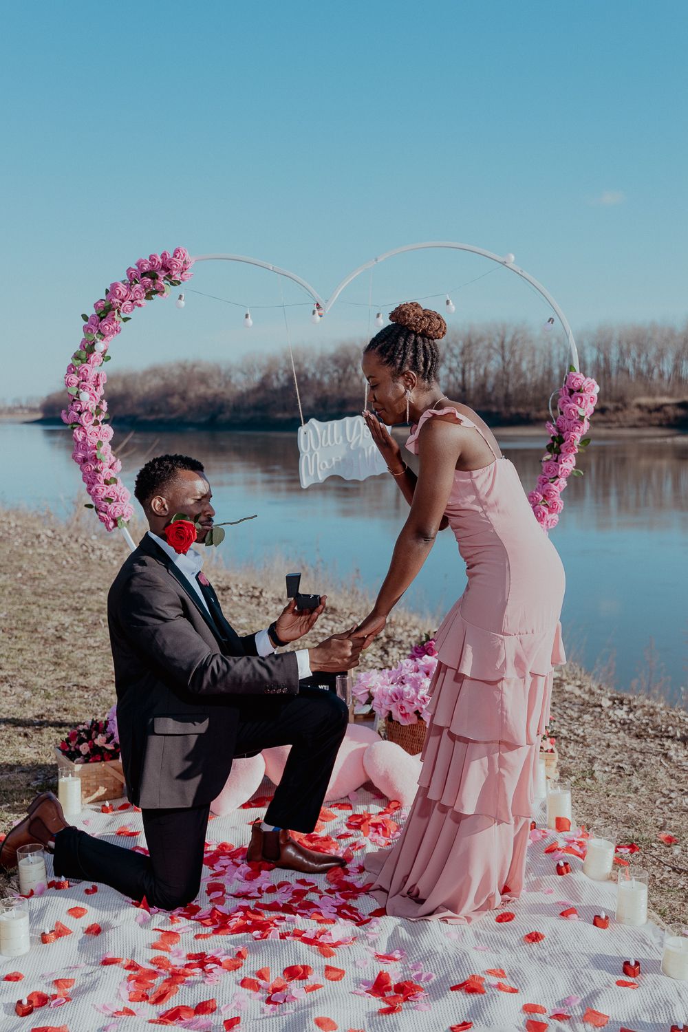 Man proposes to woman on one knee near a river, surrounded by rose petals, candles, and a heart-shaped floral arch.