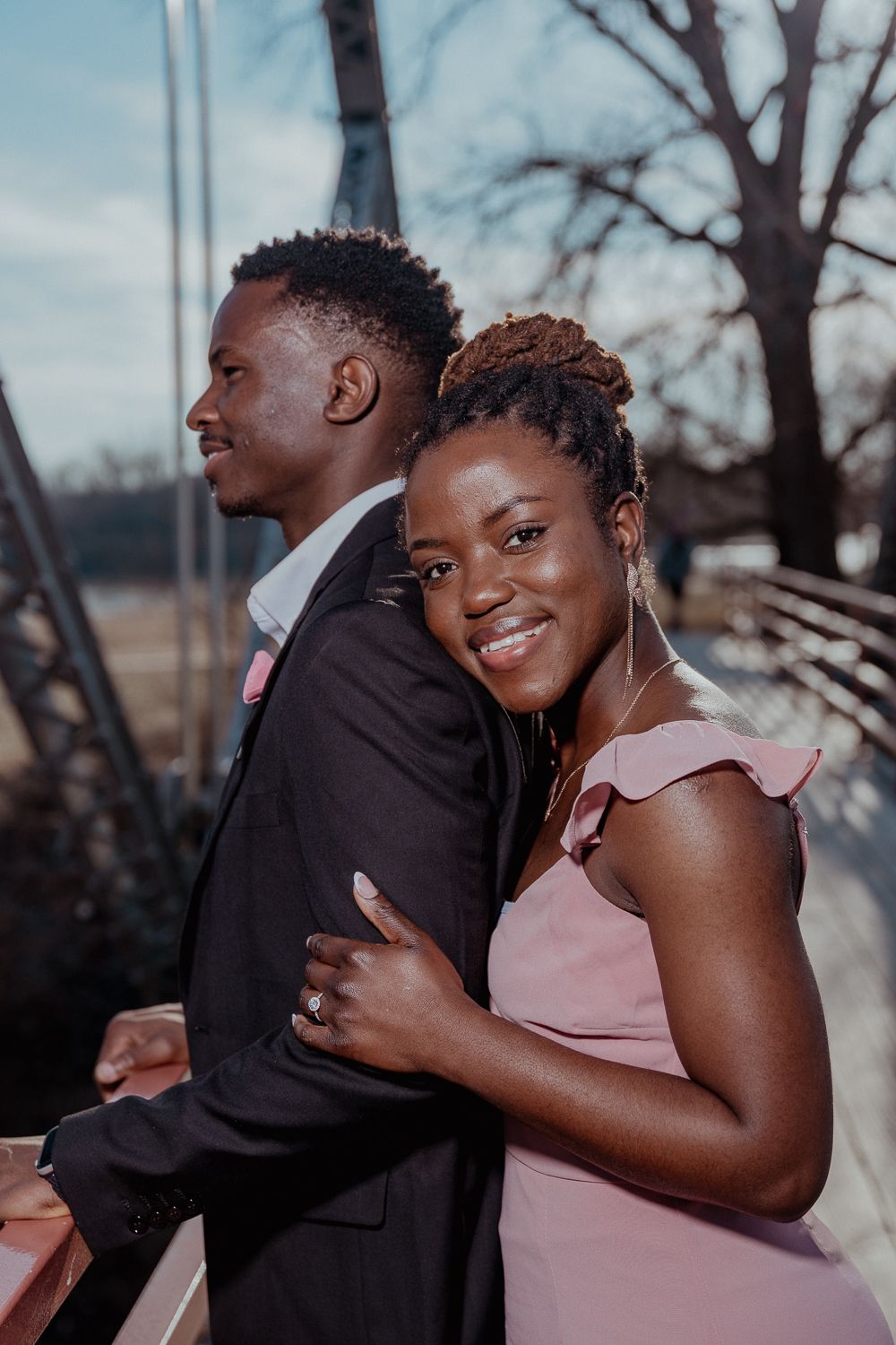 A smiling woman in a pink dress embraces a man in a black suit on a bridge during an outdoor couples photo session.
