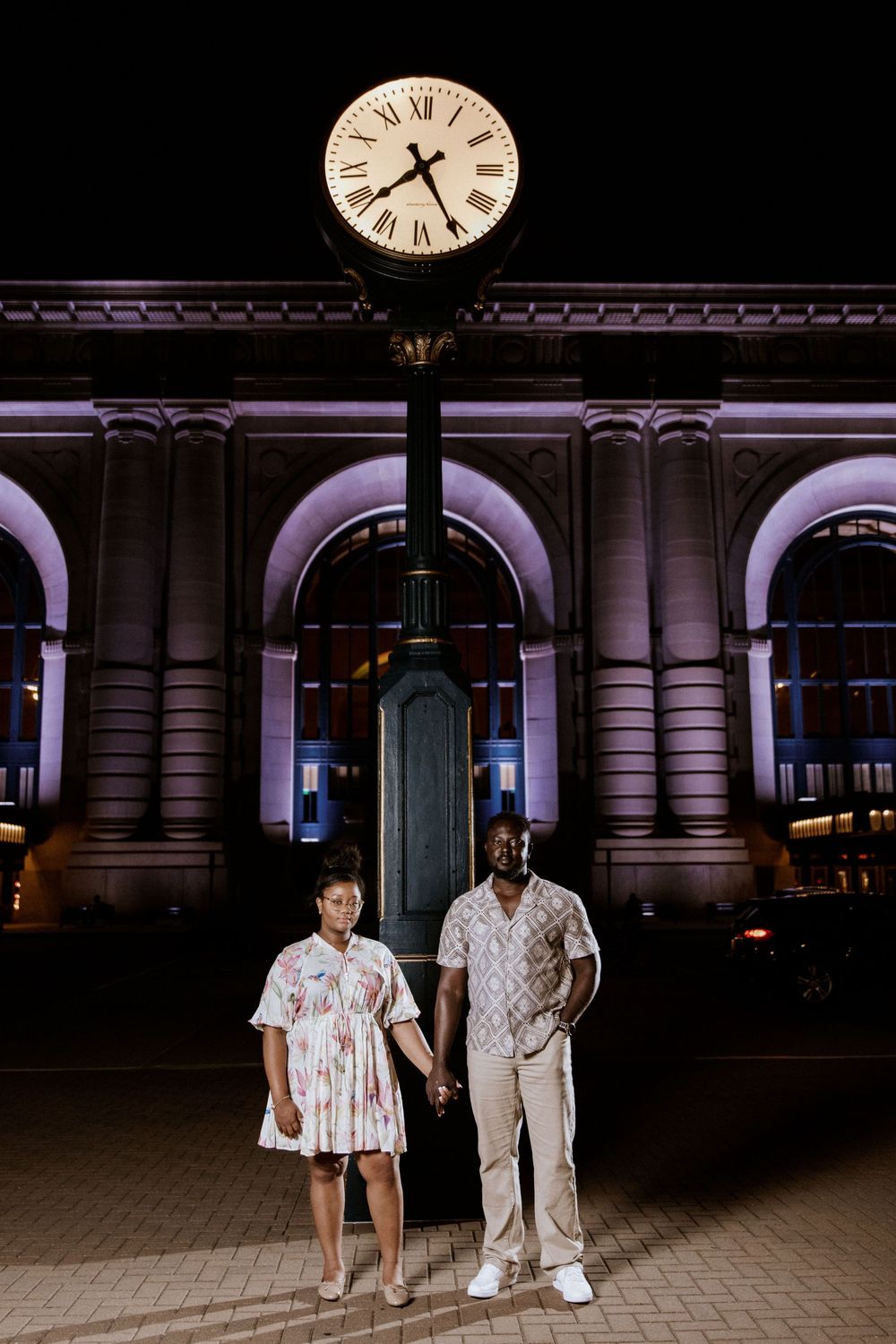 Two people stand beneath an illuminated clock in a historic train station at night.