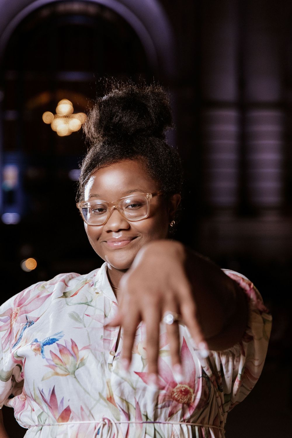 A person in glasses and a floral print dress smiles warmly in a dimly lit setting.