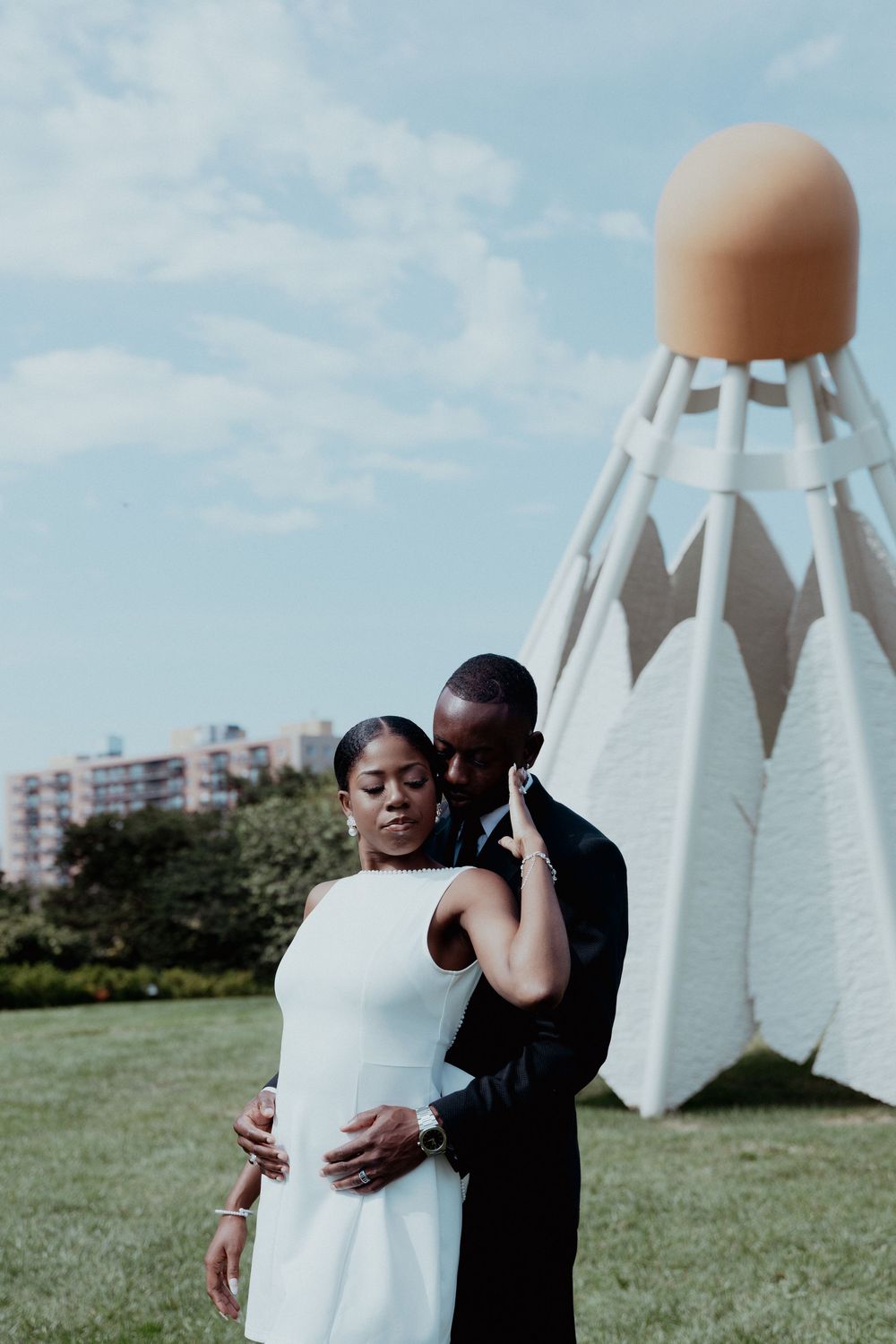 A couple embraces outdoors with white sculptural structures in the background on a grassy field.