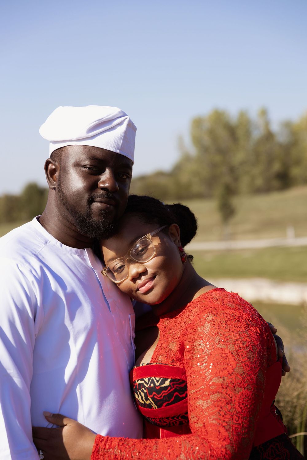 A couple embraces outdoors on a sunny day, with one person wearing white and the other in a vibrant red lace top.
