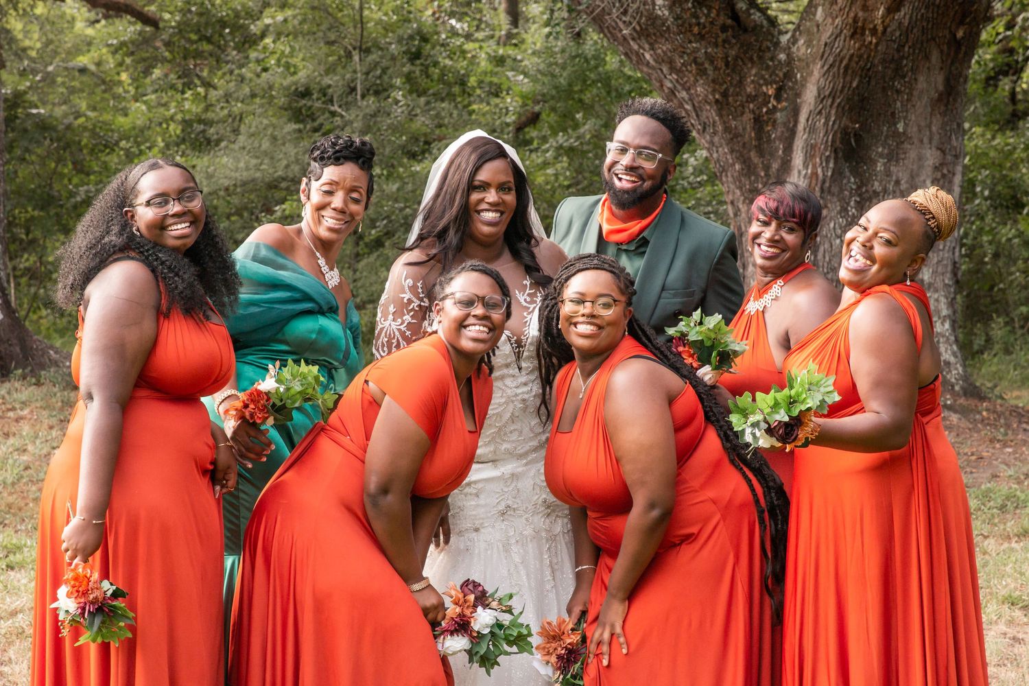 Bride and groom with bridal party in orange and green attire posing under a large oak tree.