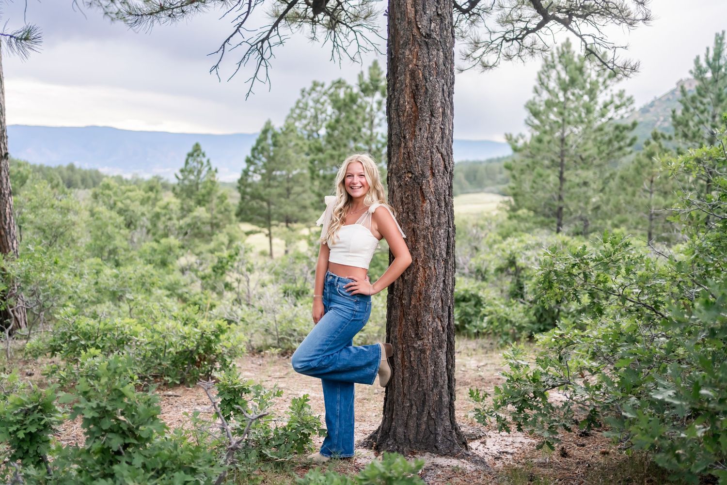 Person in athletic wear poses against pine tree with mountain landscape in background.