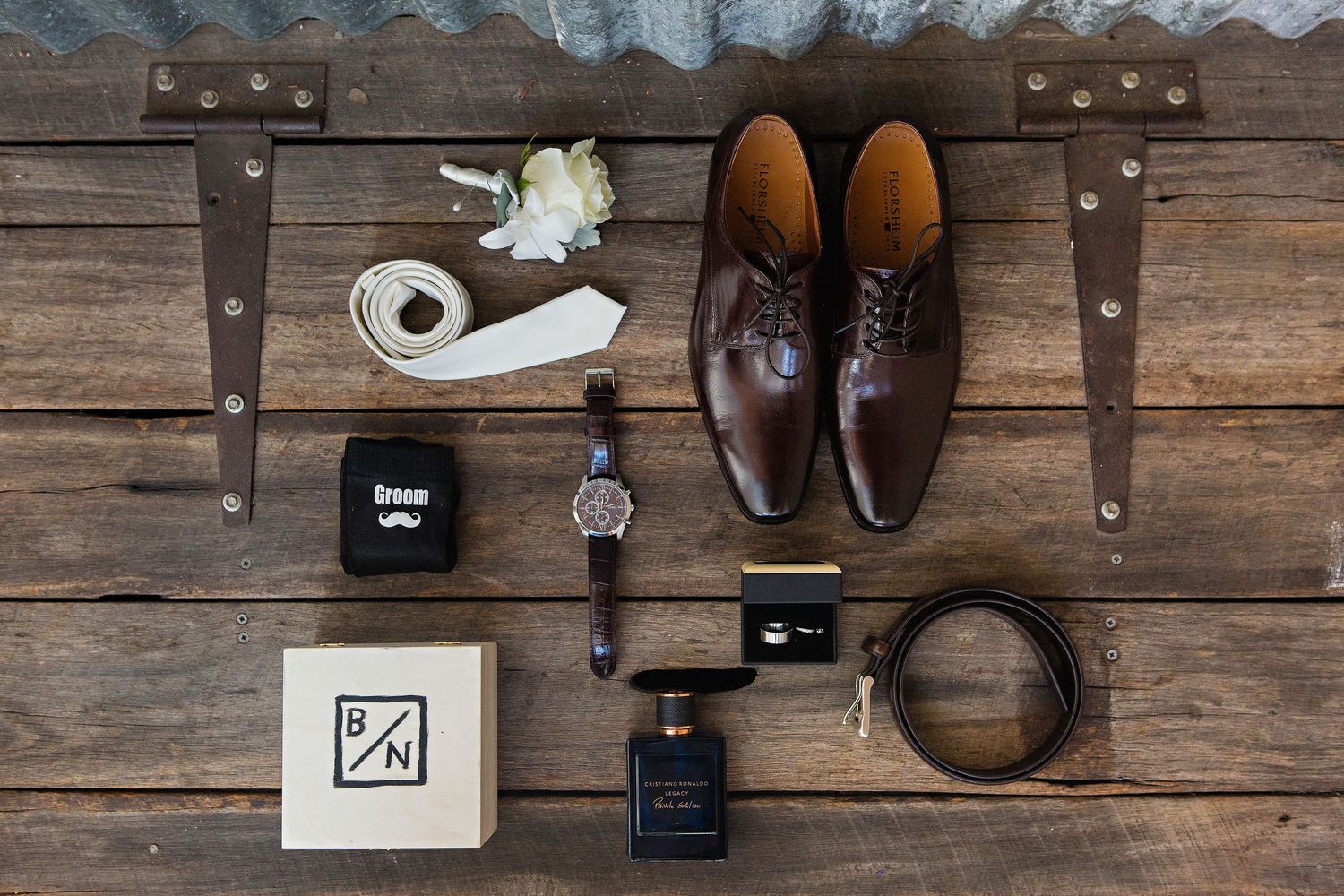 Groom prep flat lay with leather dress shoes, watch, ring box, and white tie on wooden background, by an East Anglian photographer.